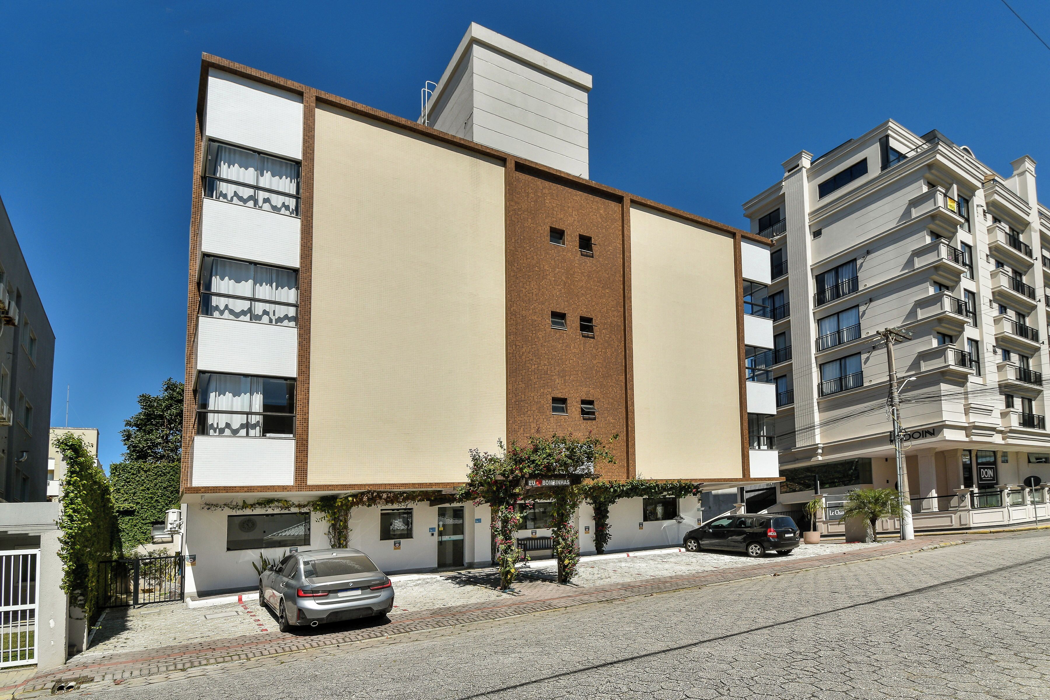 A multi-story building with a flat facade, a central entrance, and a parking area with cars.