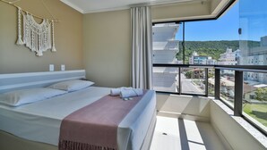 A hotel room with a bed, a macrame wall hanging, a balcony with a view of buildings and greenery. - Bella Catarina Bombinhas (Bombinhas)