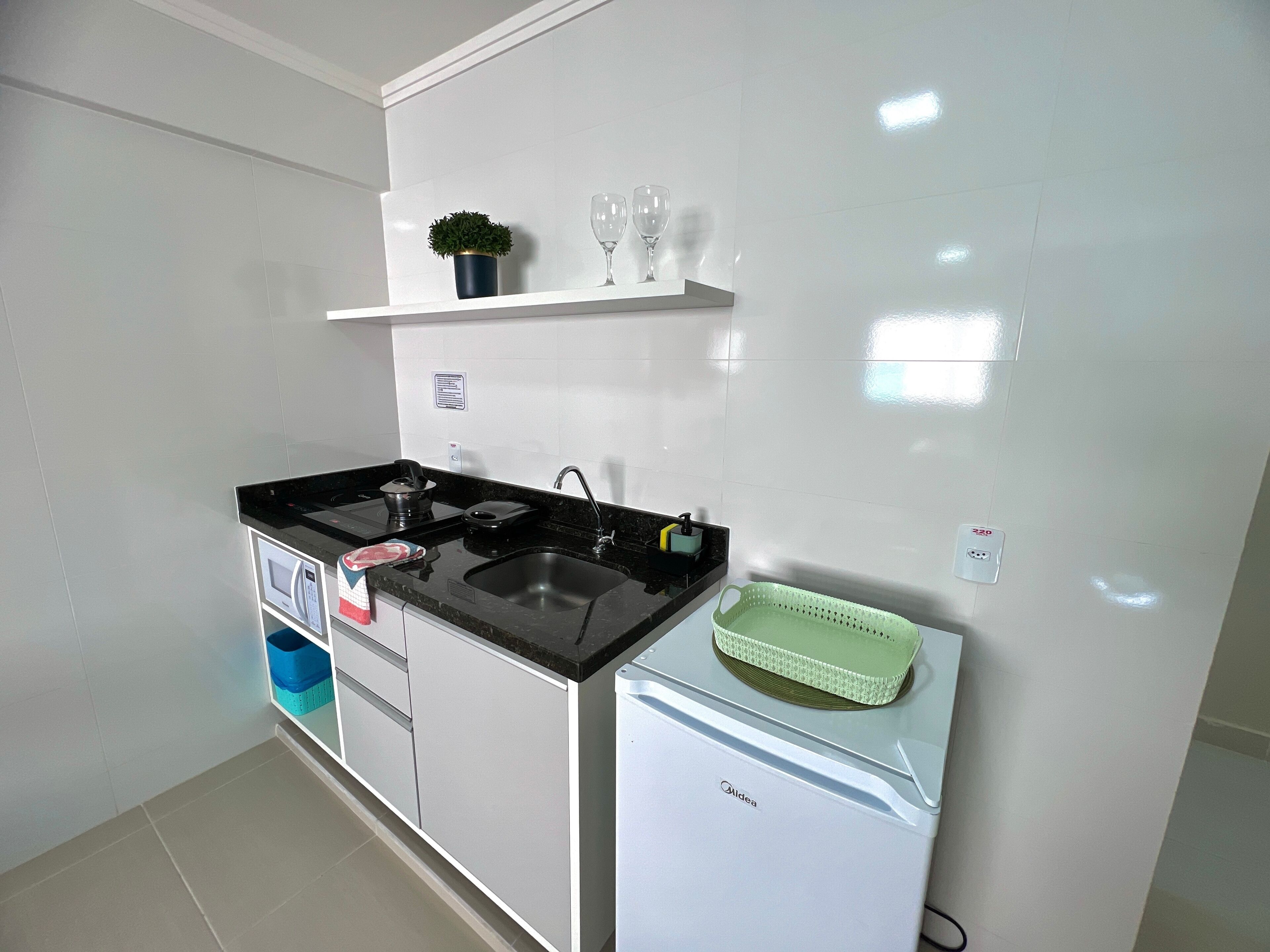 A compact kitchen with a black countertop, a white refrigerator, a sink, and a small shelf with a plant and wine glasses.