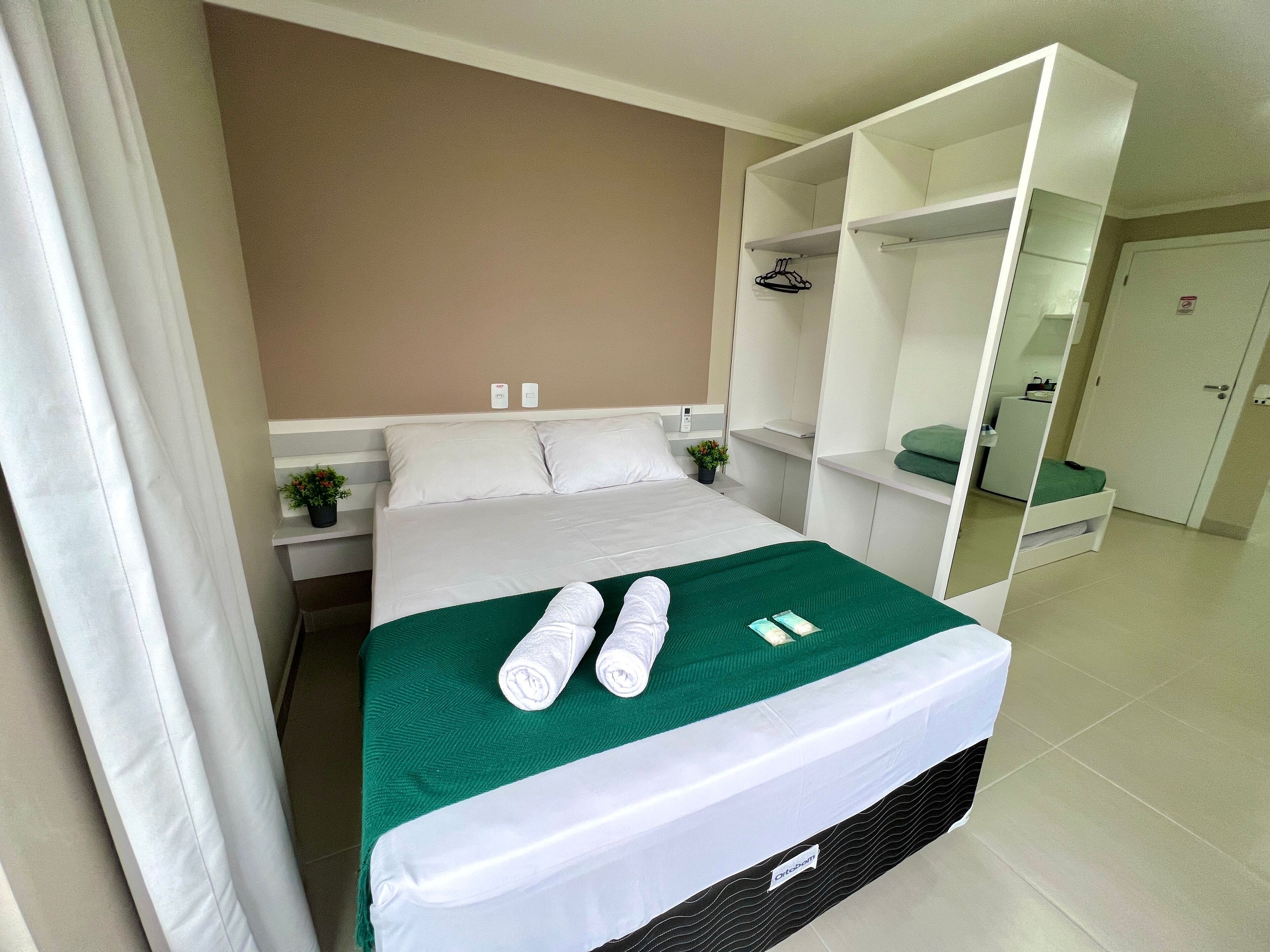 Habitación de hotel con cama, cubrecama verde, dos toallas enrolladas, cabecera blanca, mesita de noche blanca con planta y armario blanco.