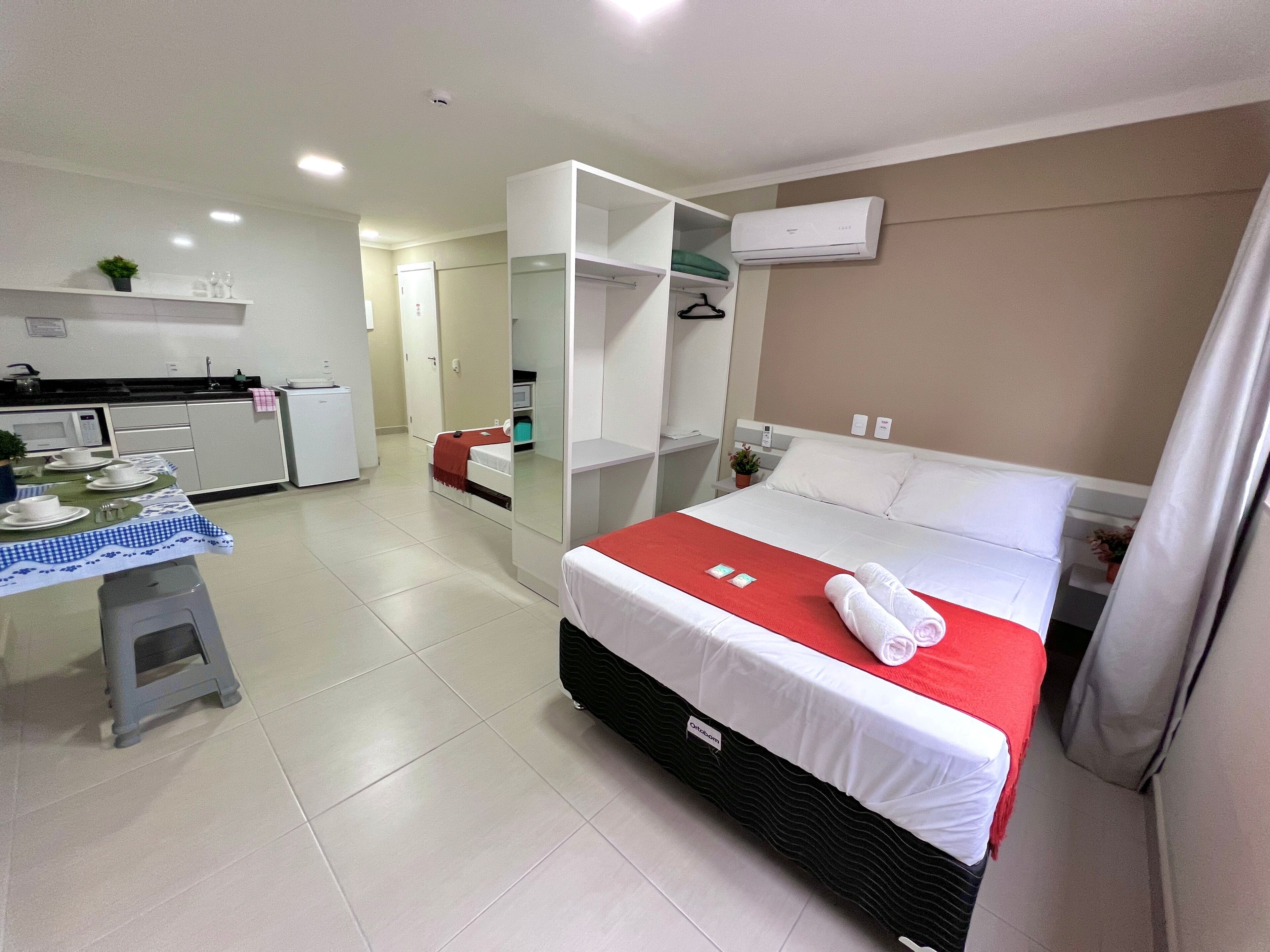 A hotel room with a bed, a small kitchenette, a mirror, and a bathroom visible through an open door.