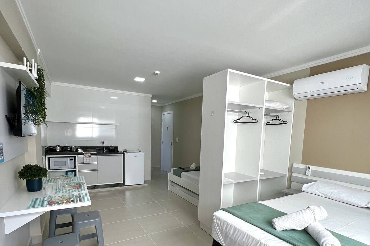 A compact hotel room with a kitchenette, a bed with a green blanket, and a wardrobe with hanging space.