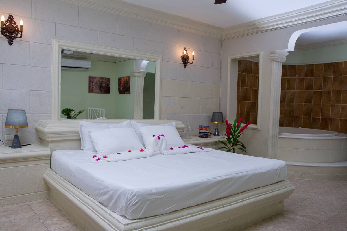 A large bed with white linens, a nightstand with a lamp, a wall-mounted light fixture, and a bathroom with a bathtub and a shower.