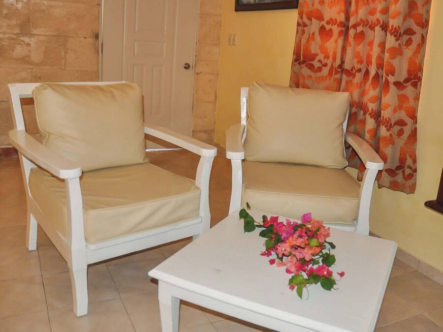 Duas poltronas brancas com almofadas bege, uma mesa branca com um arranjo floral e uma porta ao fundo.