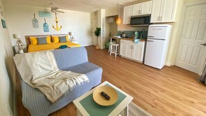 A compact living space with a sofa, a small table with a hat, a kitchen area with white cabinets and a refrigerator, and a ceiling fan. - Sea Dip Resort Studios (Daytona Beach)