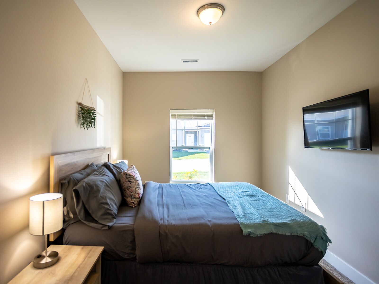 Een slaapkamer met een bed, een nachtkastje met een lamp, een aan de muur bevestigde televisie en een raam met uitzicht.