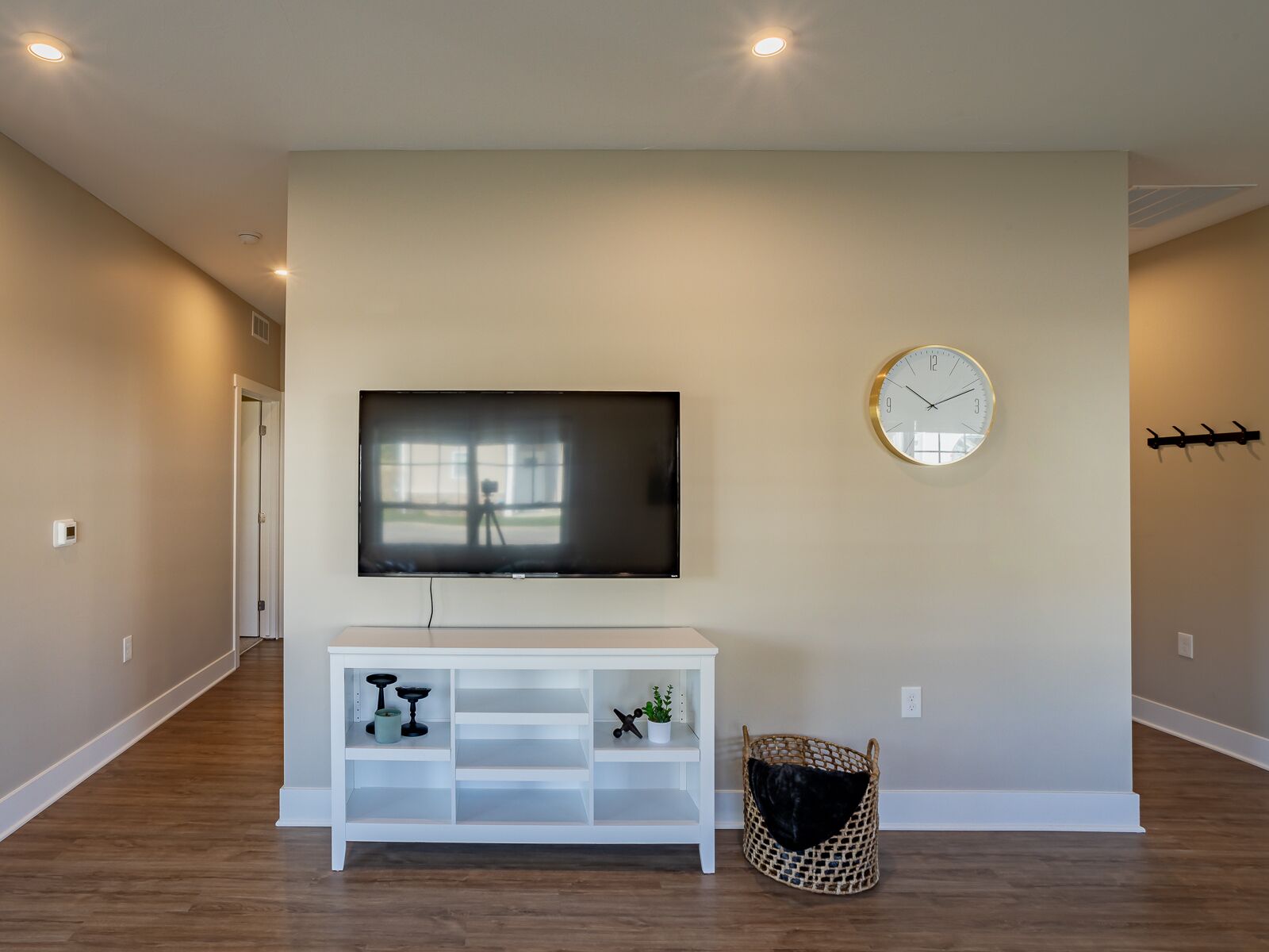 A modern living room with a flat-screen TV mounted on a white console, a round wall clock, and a woven basket with a black cushion.