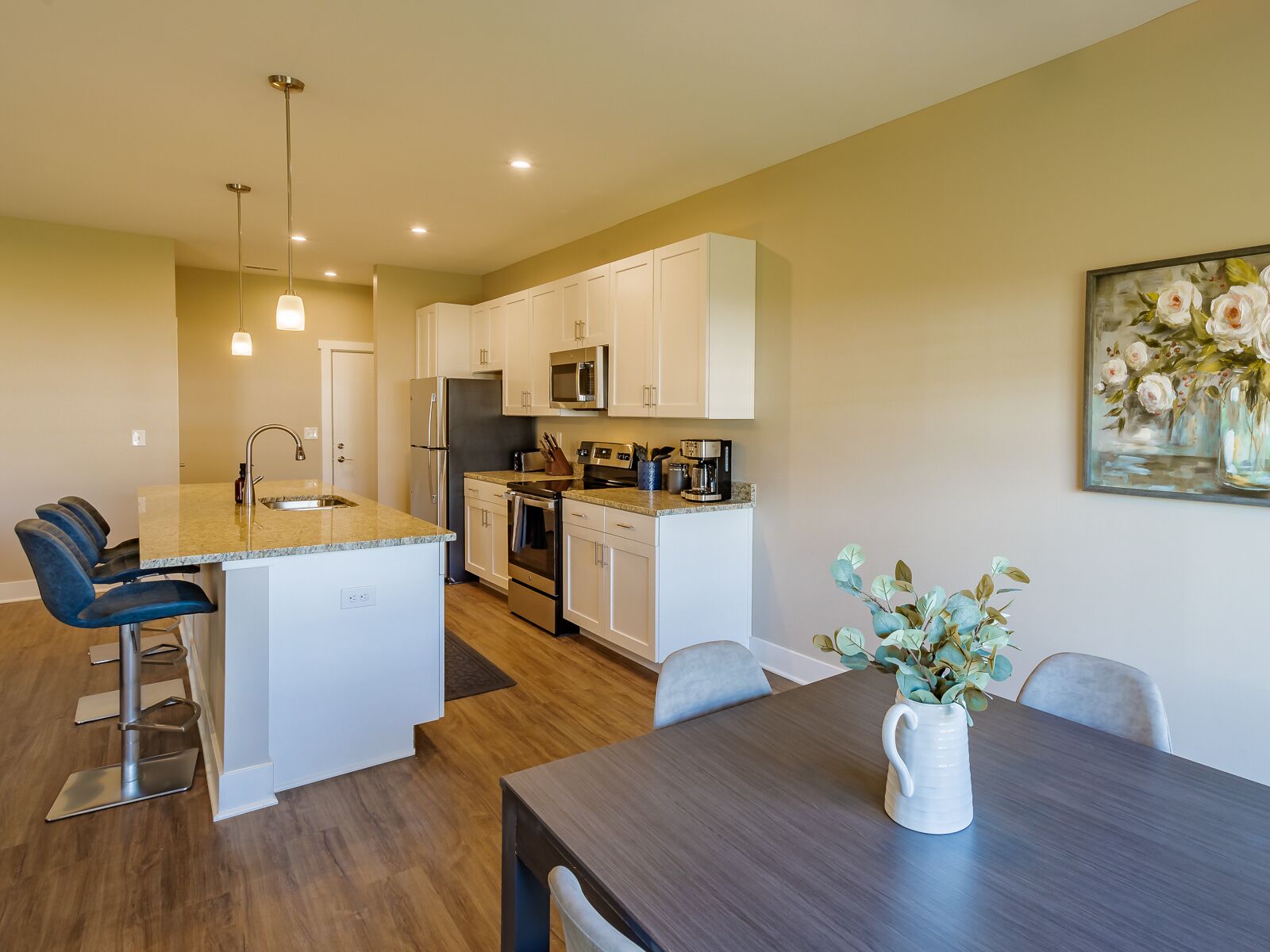 Una cocina moderna con armarios blancos, una isla central, electrodomésticos de acero inoxidable y un comedor con mesa y sillas de madera oscura.