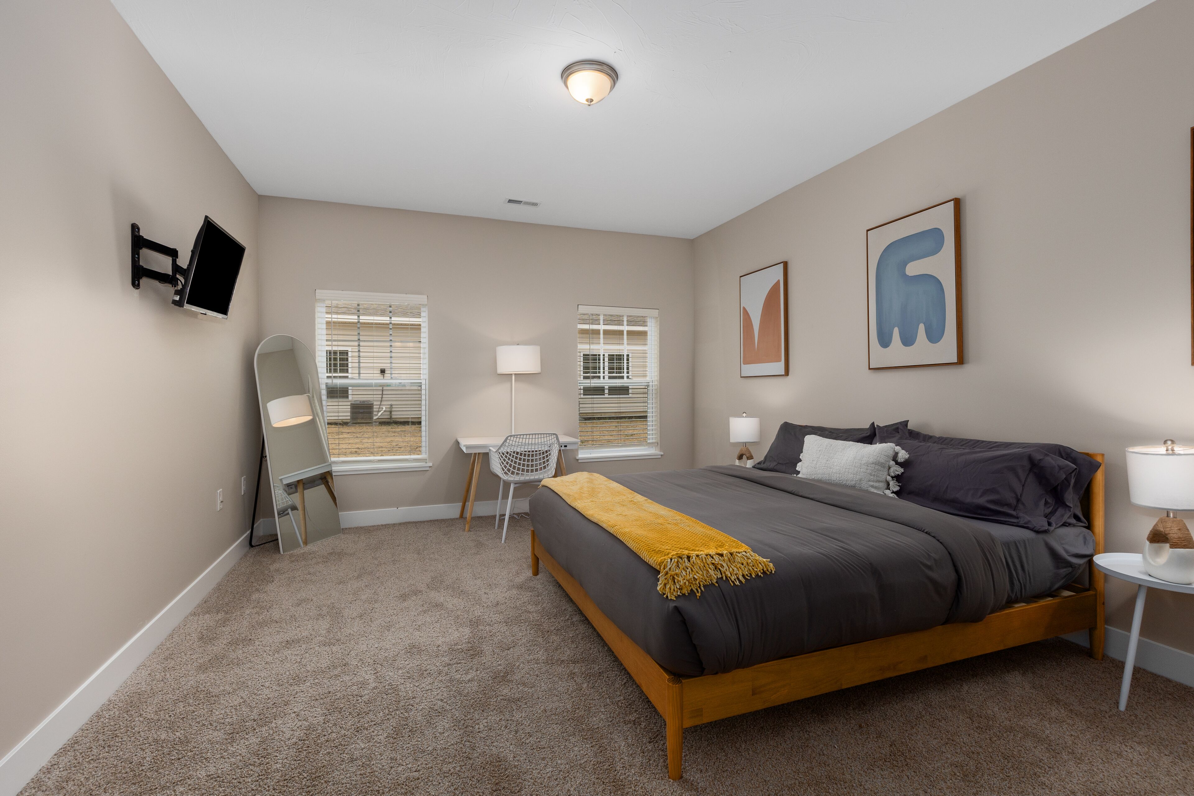 Un dormitorio con cama, escritorio, silla, dos ventanas, televisión y obras de arte en la pared.