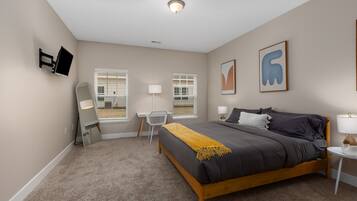 Un dormitorio con cama, escritorio, silla, dos ventanas, televisión y obras de arte en la pared.
