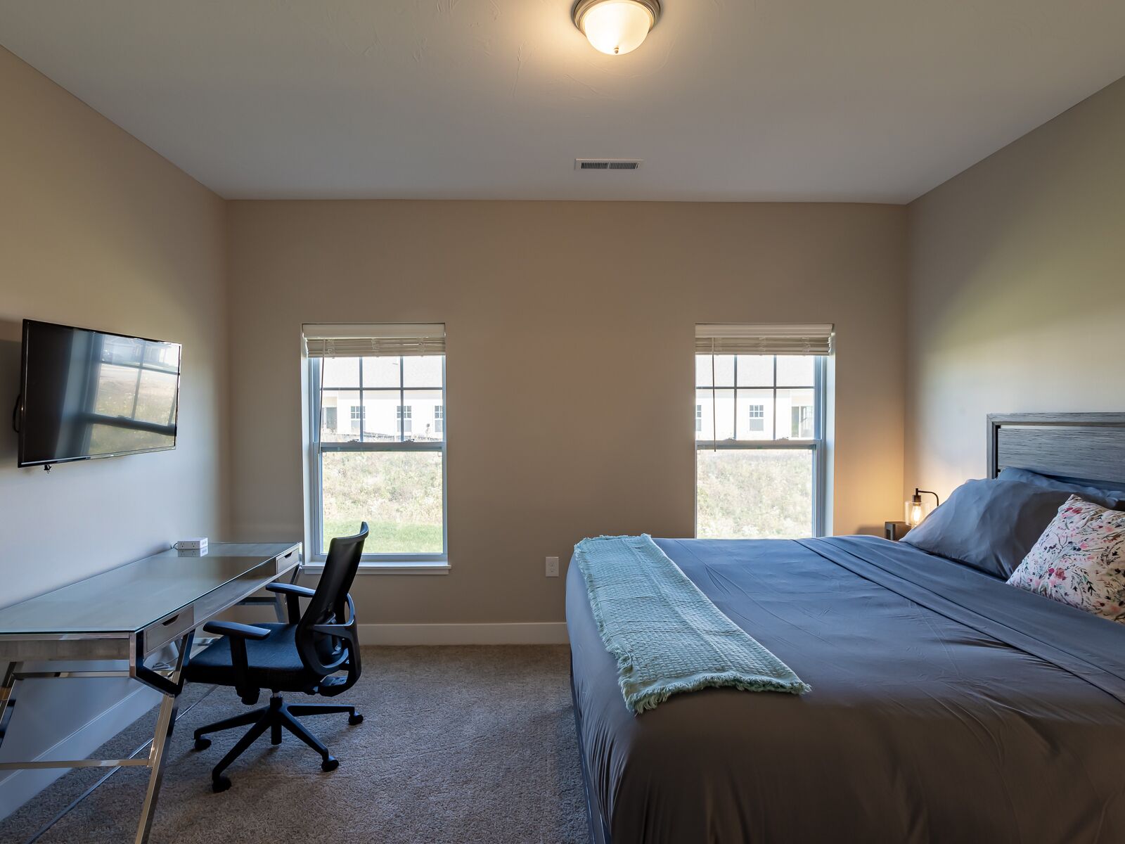 A bedroom with a bed, a desk with a chair, a TV, and two windows.