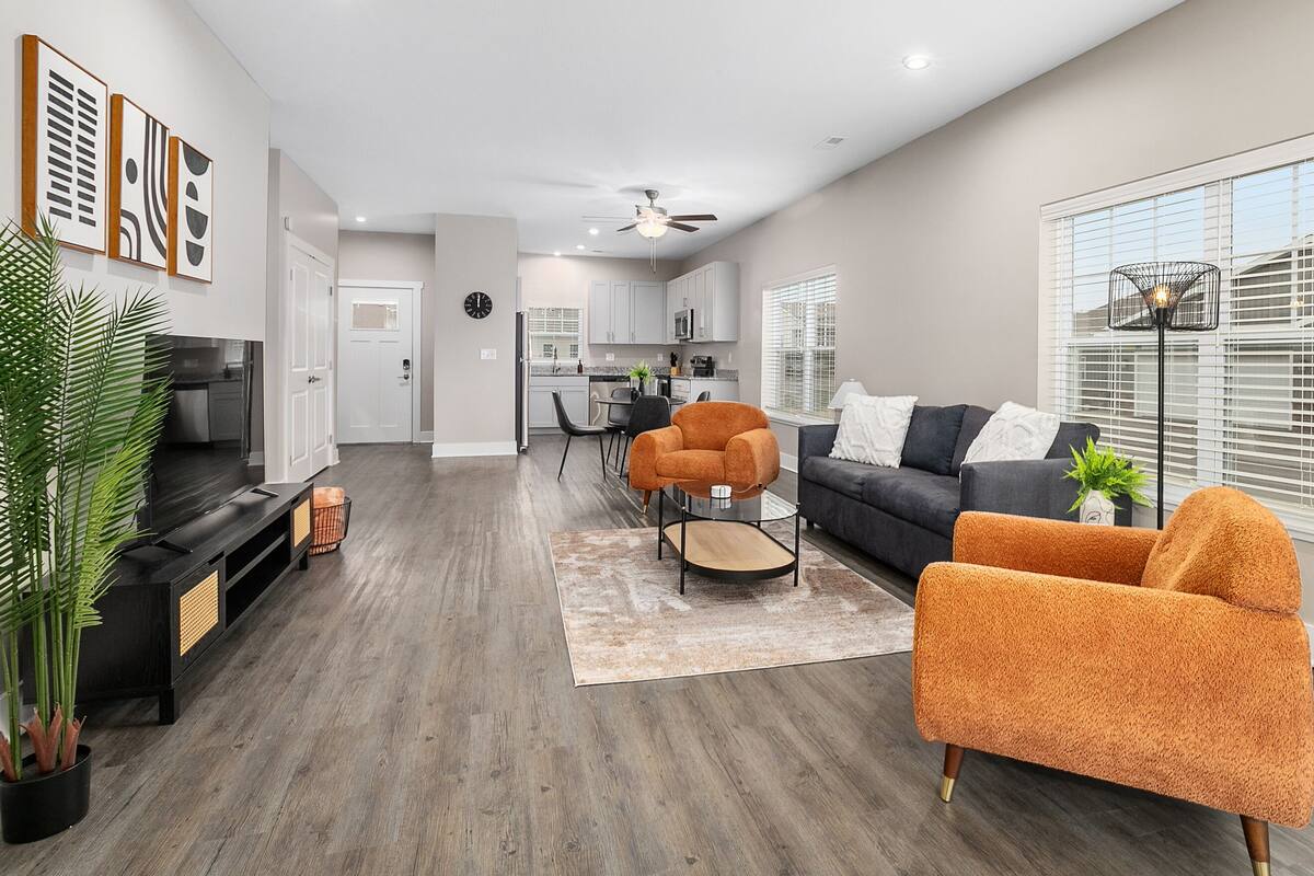 A modern living room with a sofa, armchair, and a coffee table. There is a kitchen in the background with cabinets and a microwave.