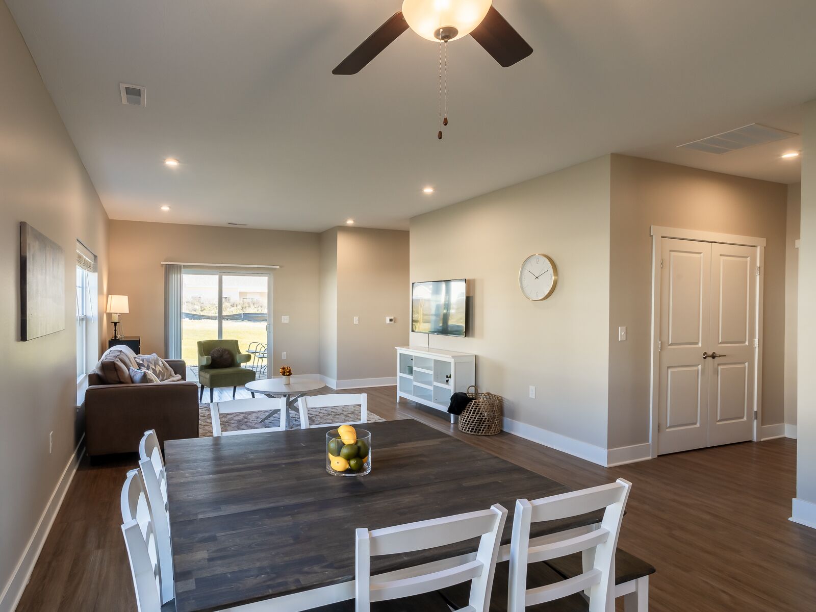 A modern living room with a dark wooden dining table, white chairs, a ceiling fan, and a large window offering a view of the outdoors.