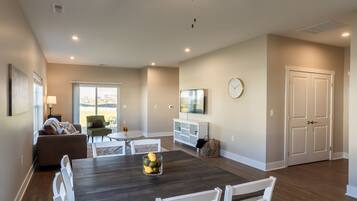 A modern living room with a dark wooden dining table, white chairs, a ceiling fan, and a large window offering a view of the outdoors.