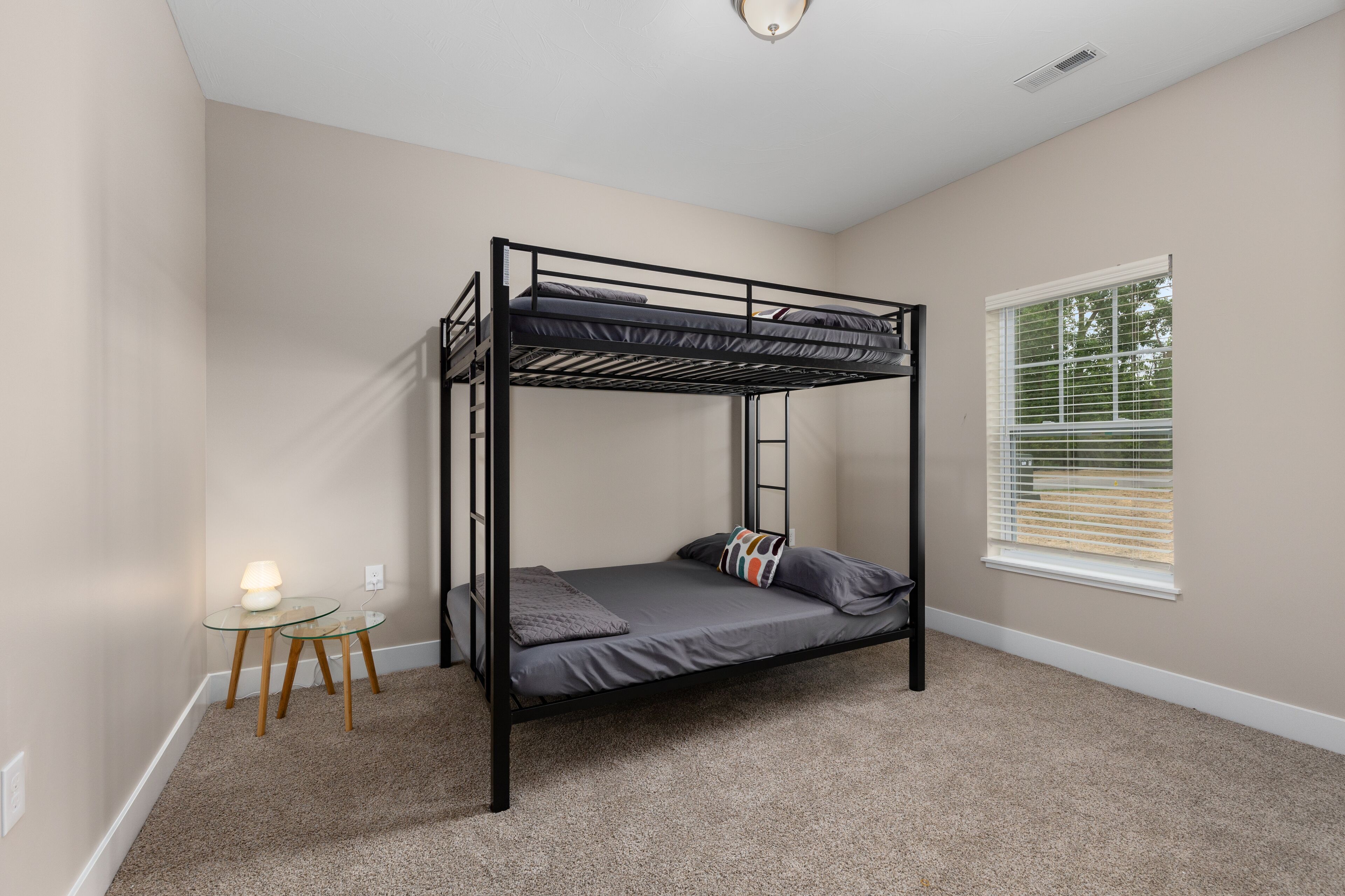 A bunk bed in a room with beige walls, a small table with a lamp, and a window with blinds.