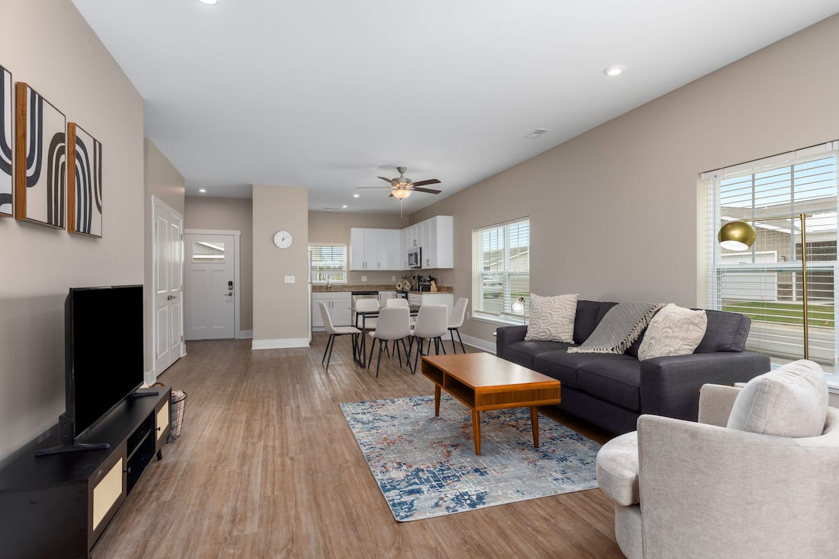 A spacious living room with a dark grey sofa, a wooden coffee table, and a TV on a black stand. There is a dining area with a white table and chairs, and a kitchen in the background.