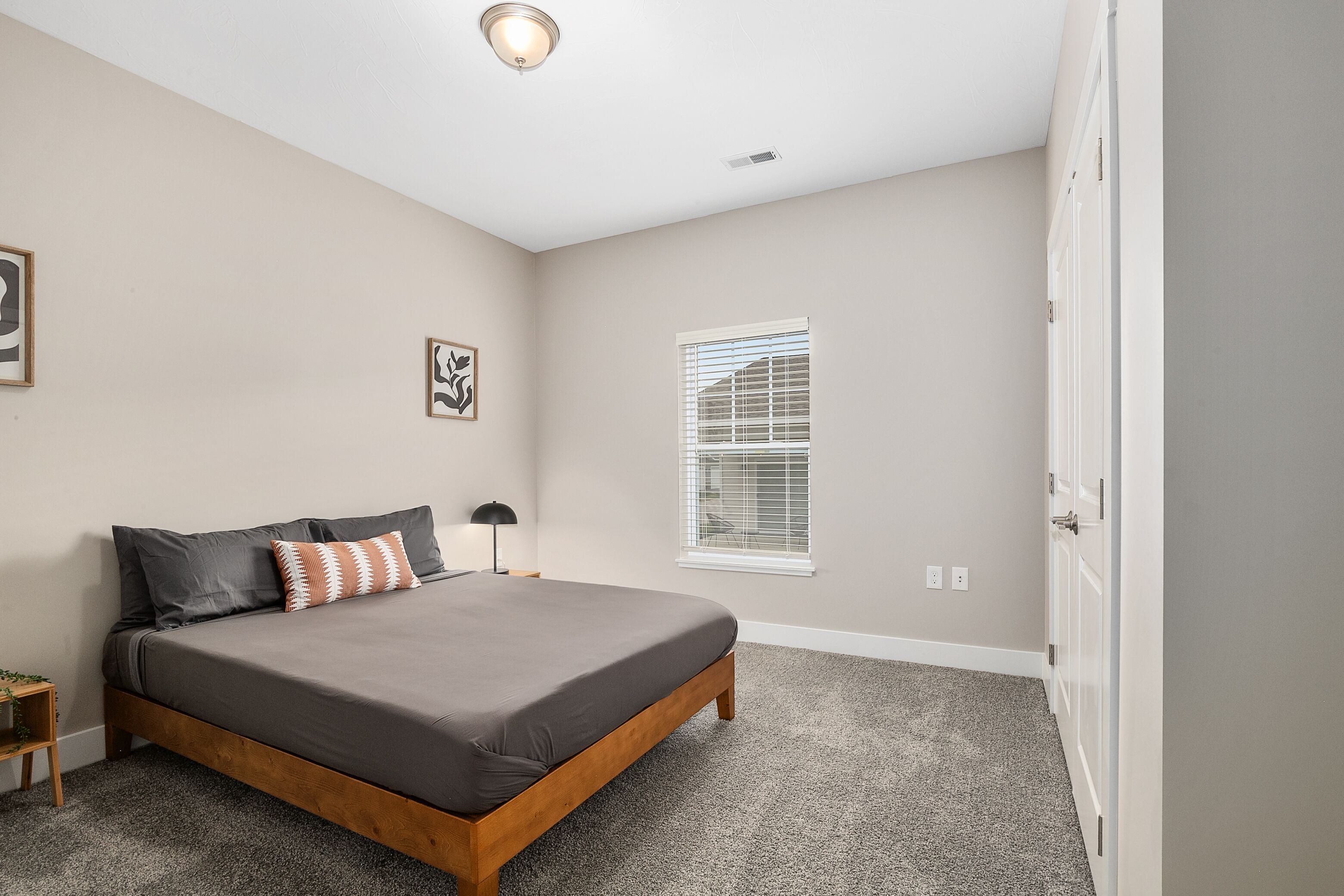 A bedroom with a bed, a nightstand, a lamp, a window with blinds, and a door.