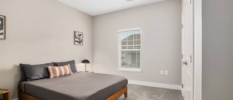 A bedroom with a bed, a nightstand, a lamp, a window with blinds, and a door.