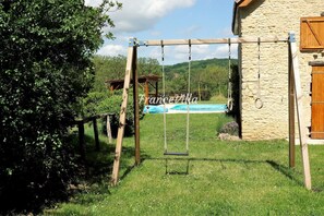 Un ensemble de balançoires en bois se trouve dans une cour verdoyante, avec un bâtiment en pierre et une piscine en arrière-plan.