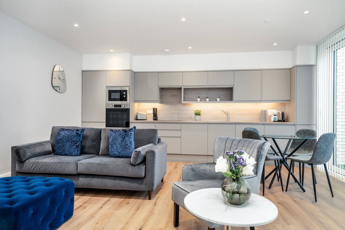 A modern living room with a grey sofa, blue ottoman, and a dining area with a glass table and chairs.