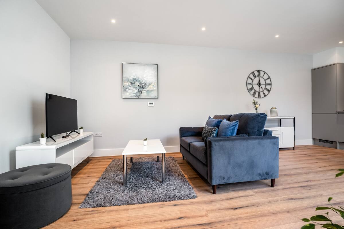 A modern living room with a flat-screen TV, a grey sofa, a coffee table, and a large wall clock.