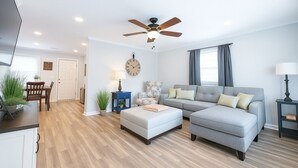 Un salon moderne avec un canapé d’angle gris, un pouf blanc, une table d’appoint bleue, un plancher en bois et un ventilateur de plafond.