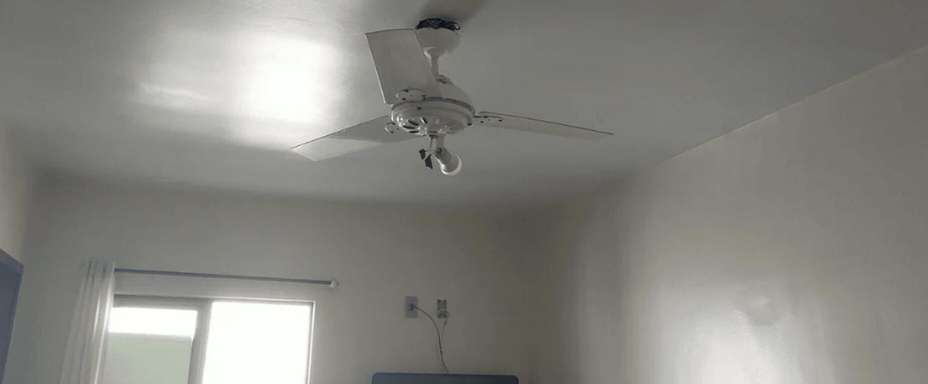 Una ventola a soffitto in una stanza con pareti bianche e una finestra.