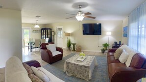 Una sala de estar con ventilador de techo, televisor, comedor con sillas y una mesa, y vista al exterior.