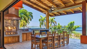 Cozinha externa com churrasqueira, mesa de jantar e cadeiras, com vista para uma praia com palmeiras.