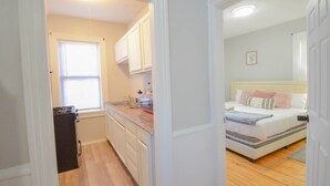 A compact room with a kitchenette, a bed with a headboard, and a window with blinds.