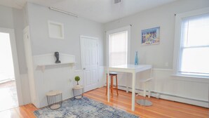 A room with wooden flooring, a white door, a white desk with a chair, a blue rug, and a small plant on a side table.