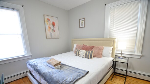 A bedroom with a bed, a bedside table, a window, and a wall with two framed pictures.