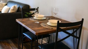 Un comedor con una mesa de madera dispuesta para dos personas, dos sillas negras, un sofá negro con cojines, un cuadro pequeño en la pared y un termostato.