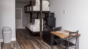 Una habitación pequeña con una cama litera, un refrigerador compacto, un microondas, una mesa de madera con sillas y un cubo de basura.