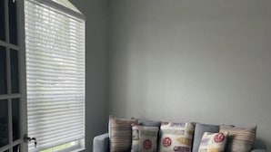 Una habitación con un sofá gris, cojines estampados, un ventilador de techo y una ventana con persianas.