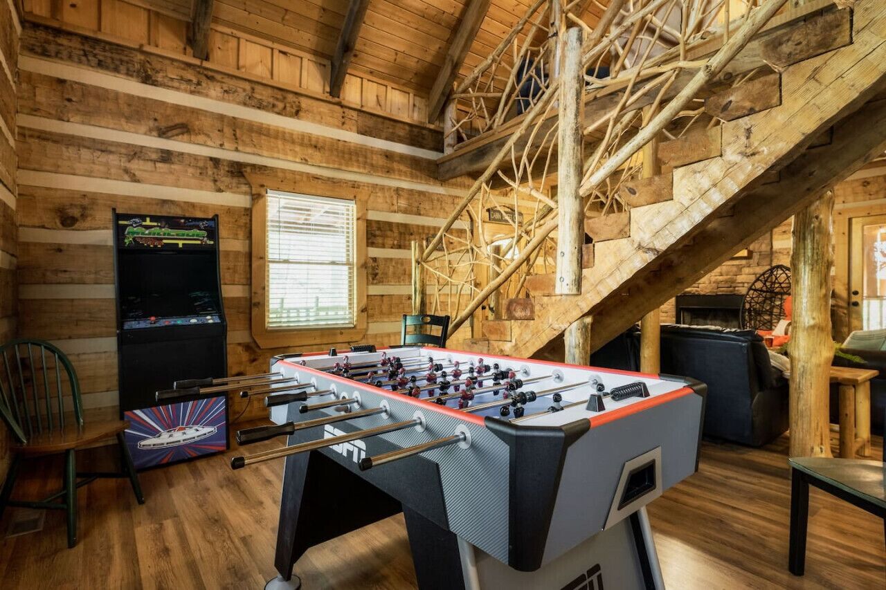A rustic wooden interior with a foosball table, a vintage arcade machine, and a staircase.