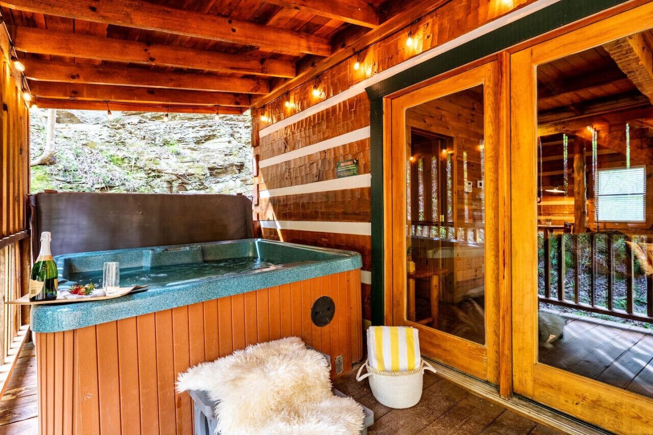A wooden cabin with a hot tub, a wooden deck, and a stone wall in the background.