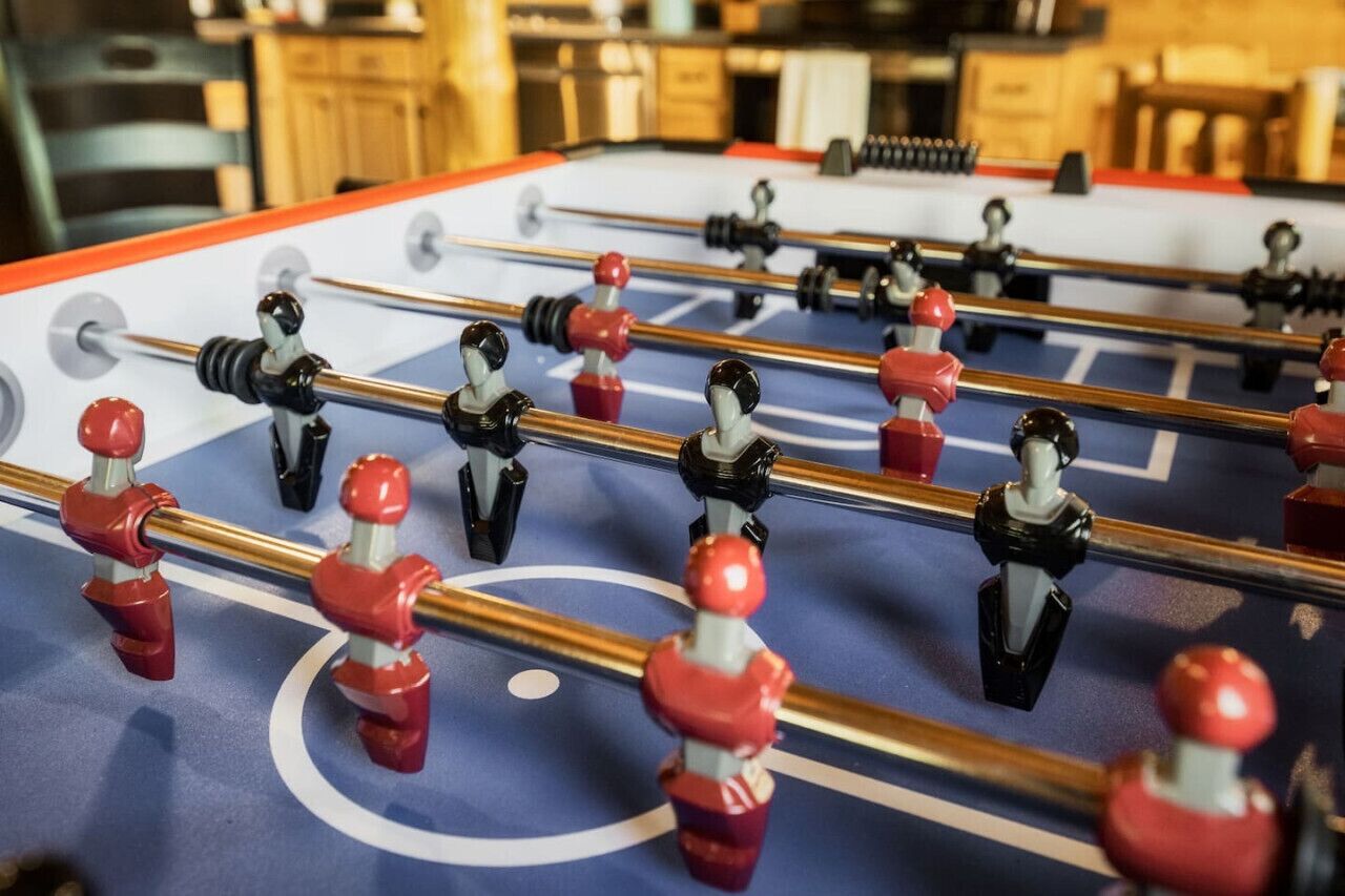 Une table de baby-foot avec des joueurs rouges et noirs, dans une pièce dotée de meubles en bois et recouverte d’une nappe bleue.