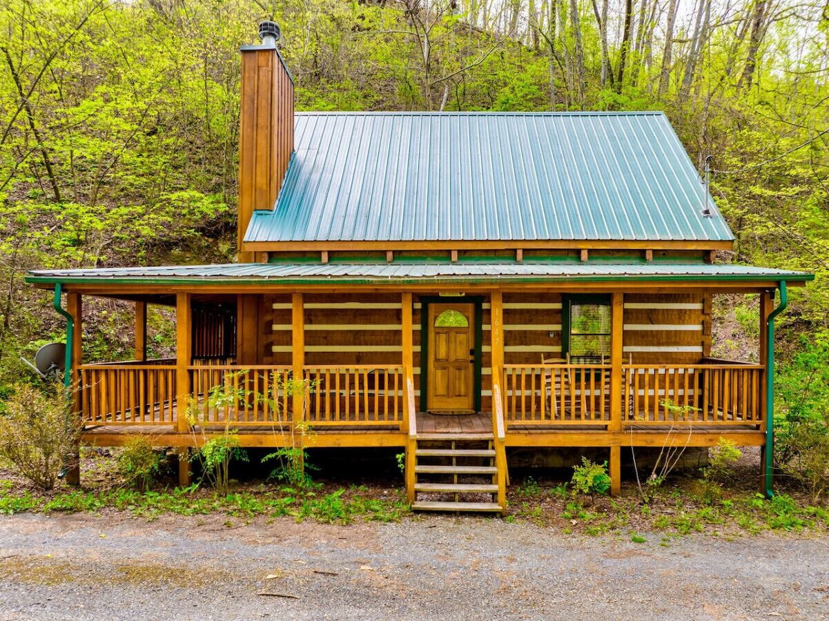 Une cabane en rondins avec un toit en métal vert, un revêtement extérieur en bois et une véranda à l’avant, dotée d’une porte en bois.