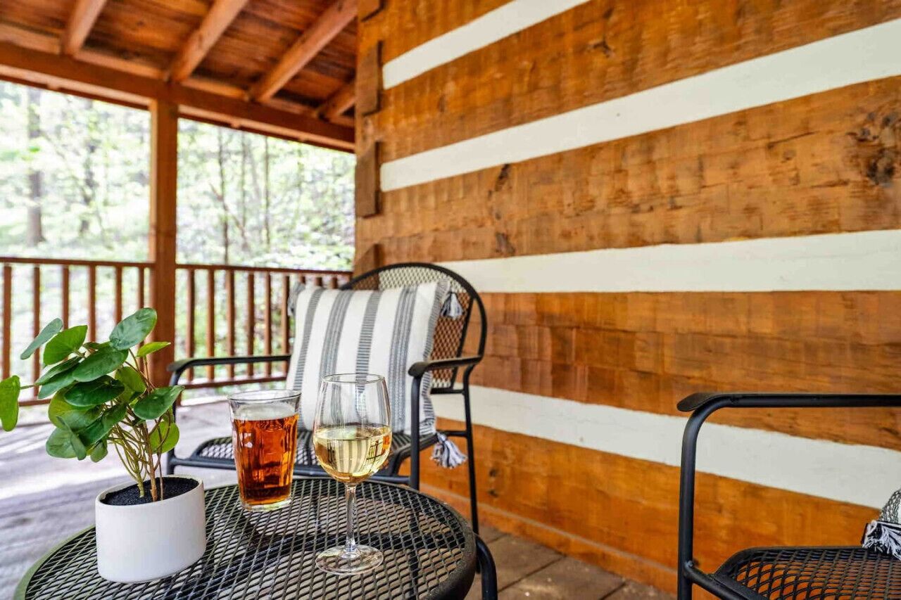 A covered outdoor seating area with a wooden wall, a metal chair, a cushioned chair, a glass of white wine, and a potted plant.