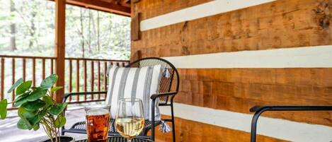 A covered outdoor seating area with a wooden wall, a metal chair, a cushioned chair, a glass of white wine, and a potted plant.