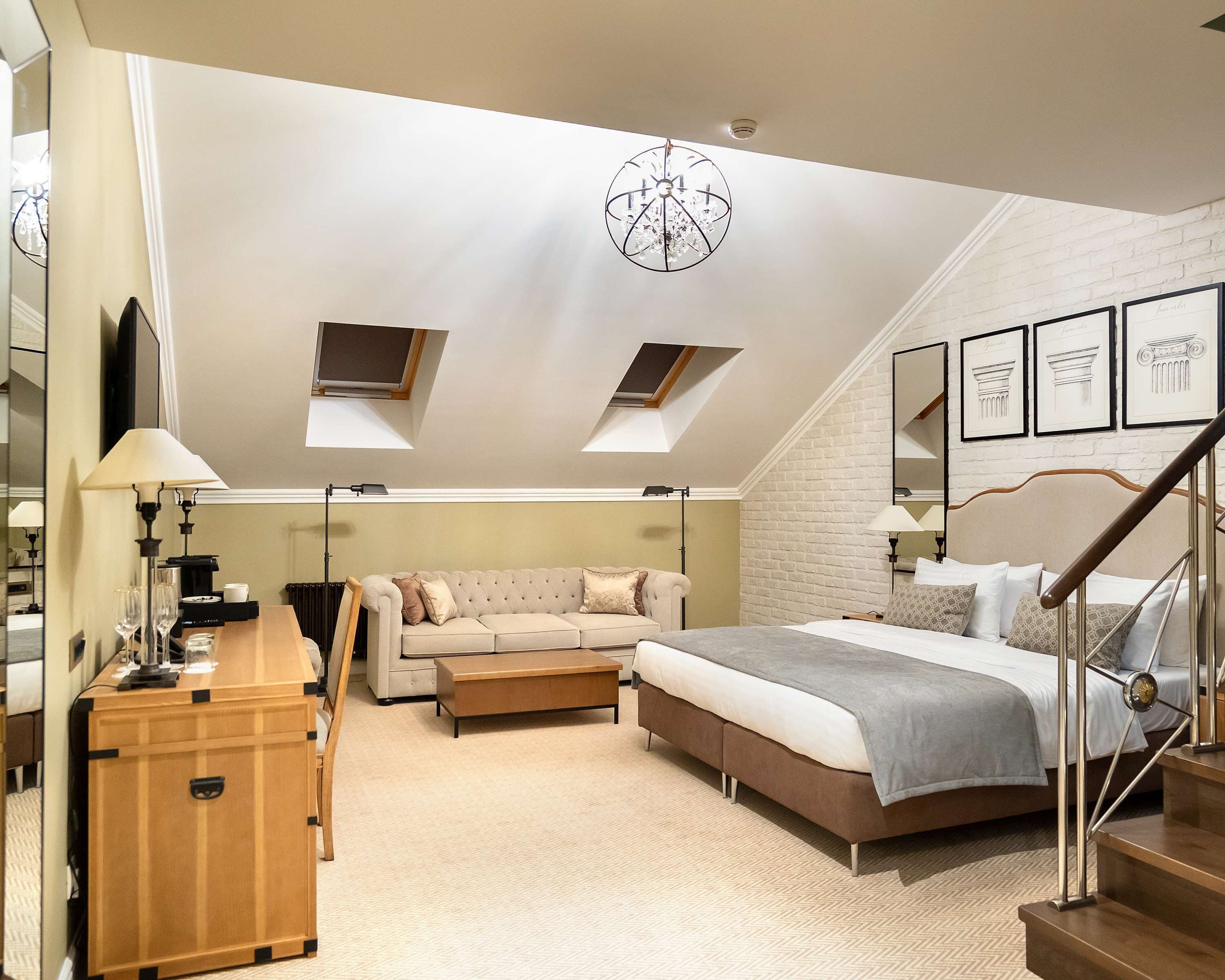 A bedroom with a large bed, a sofa, a desk, and a chest of drawers. There are skylights on the ceiling and a staircase leading to an upper level.