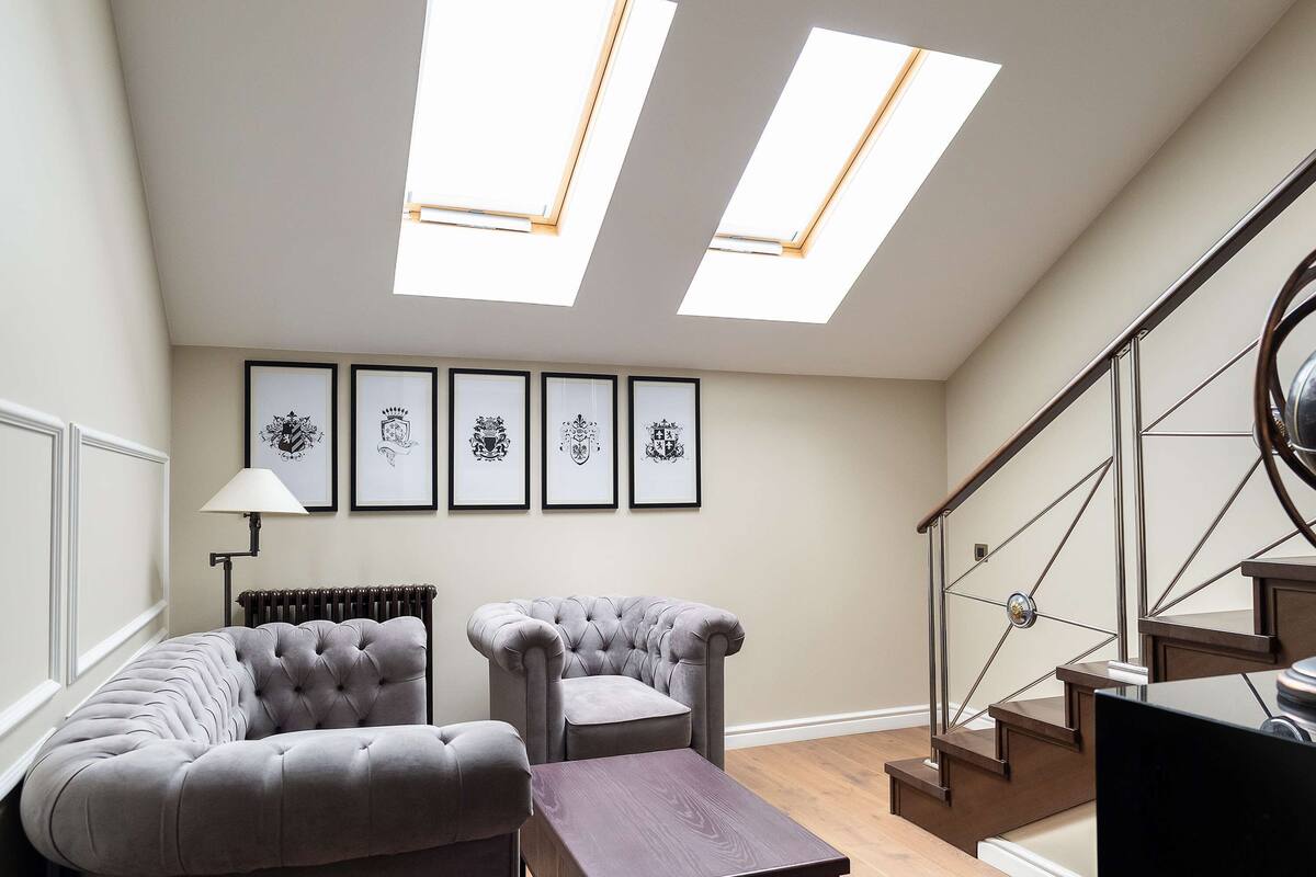 A spacious living room with a high ceiling, two skylights, a staircase, and a series of framed heraldic shields on the wall.