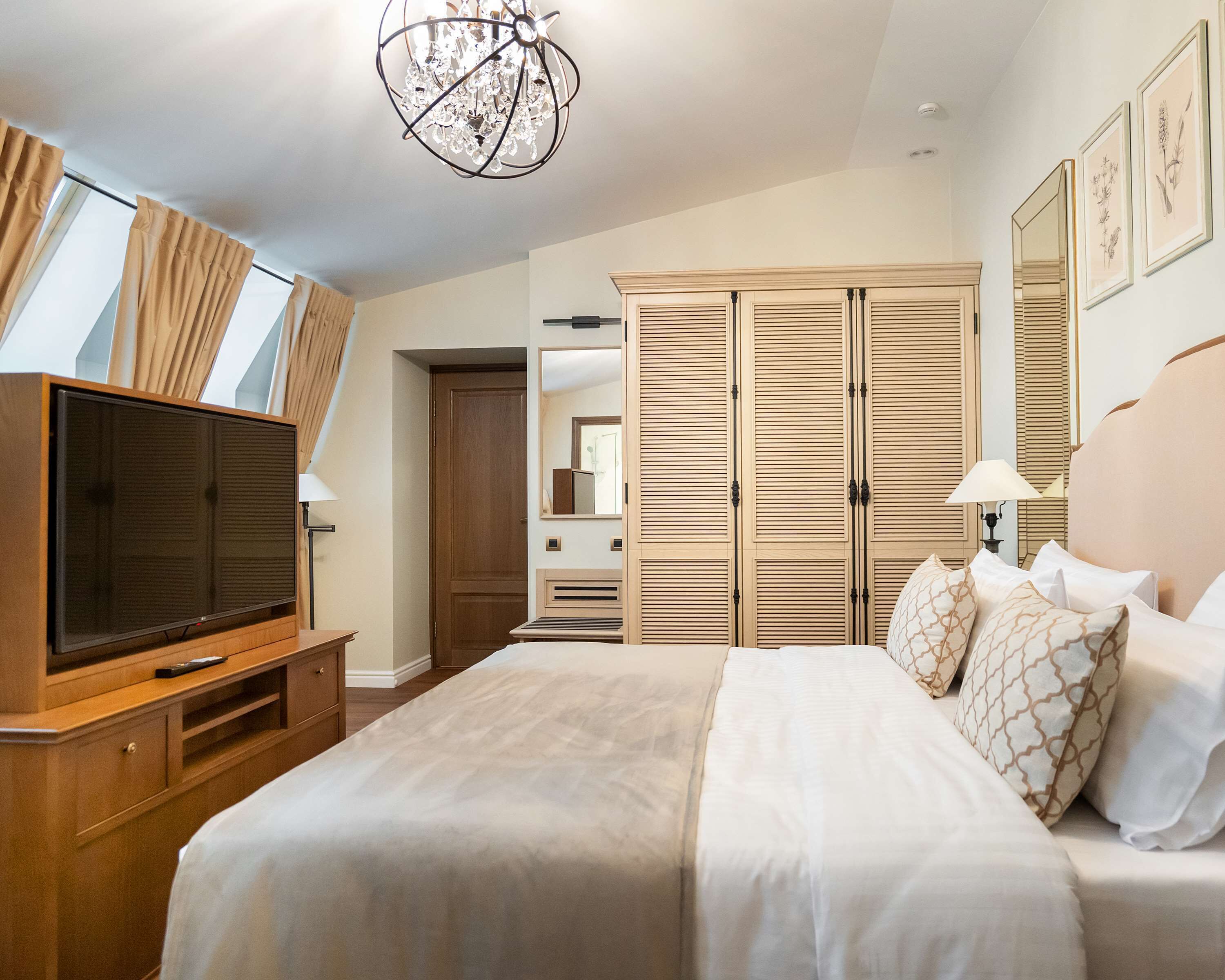 A hotel room with a large bed, a television, a wardrobe, and a chandelier.