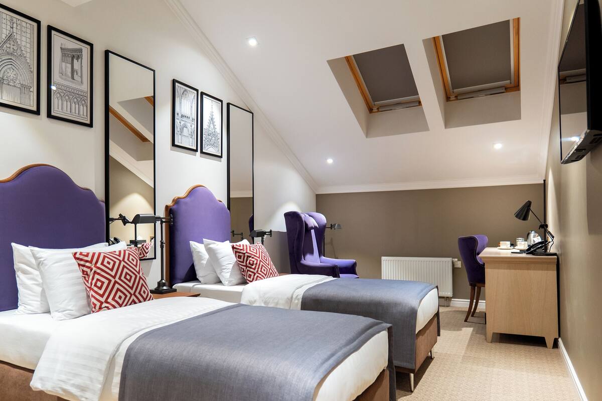 A bedroom with two beds, a desk, and a chair. There are framed pictures on the wall and a skylight above the beds.