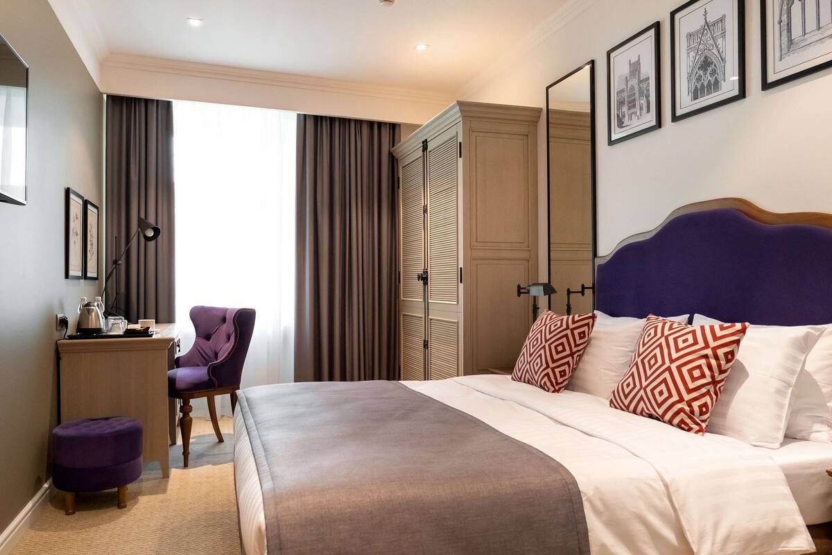 A hotel room with a large bed, a desk with a chair, a wardrobe, and two framed pictures on the wall.