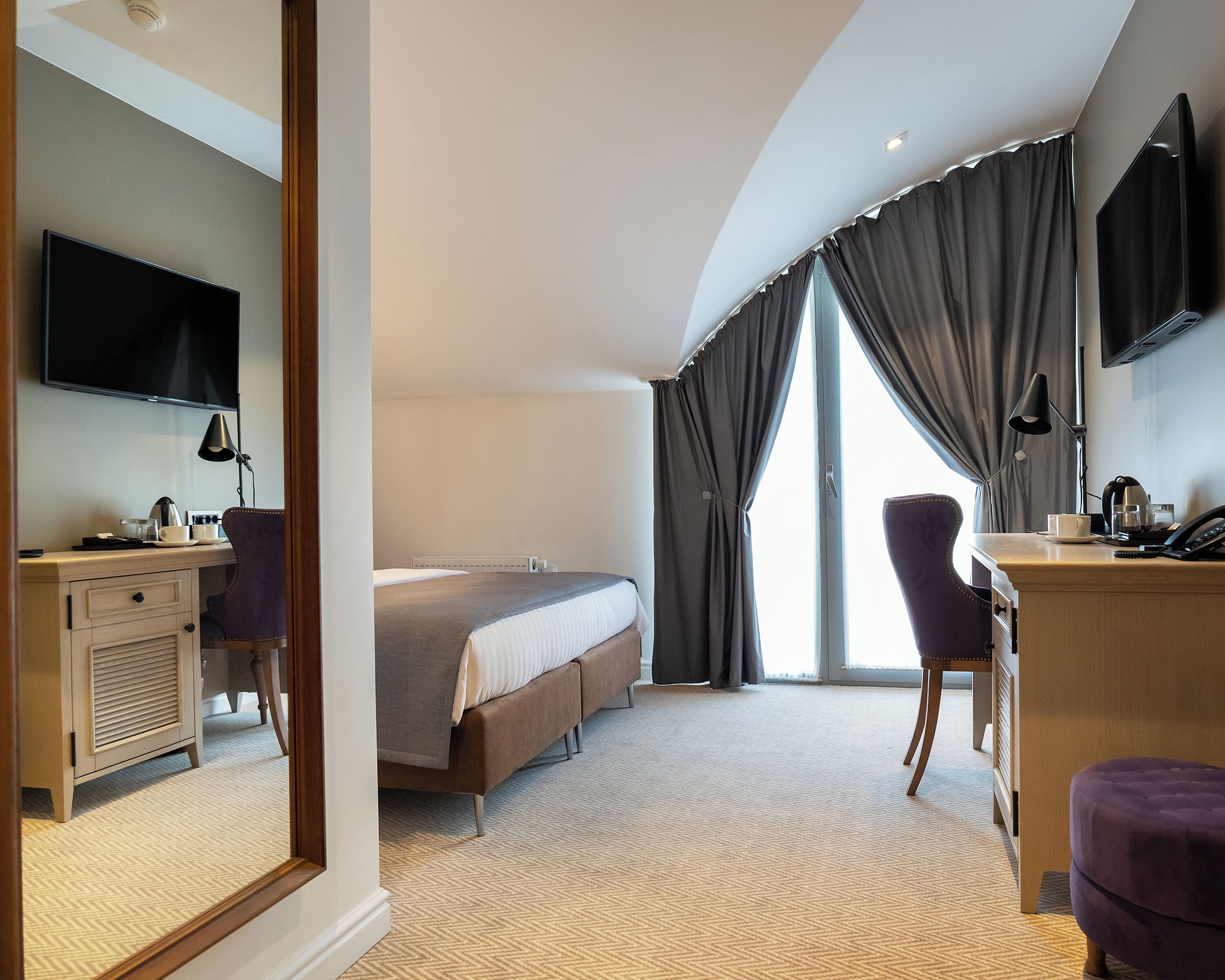 A hotel room with a bed, a desk with a computer, a chair, a TV mounted on the wall, and a large window with curtains.
