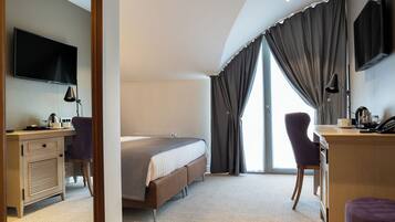 A hotel room with a bed, a desk with a computer, a chair, a TV mounted on the wall, and a large window with curtains.