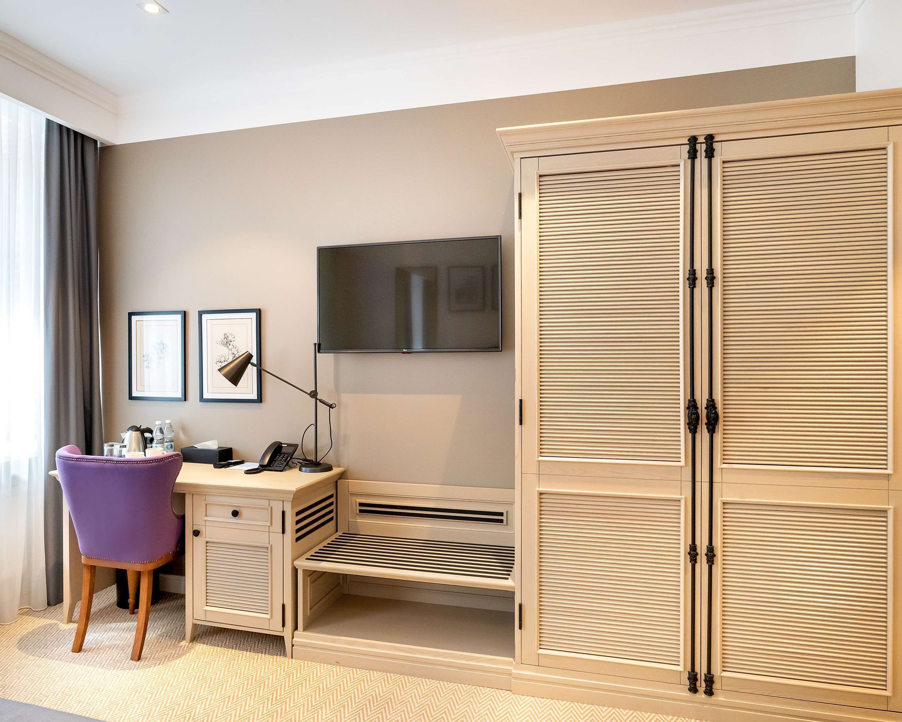 Une chambre d’hôtel dotée d’une grande armoire, d’un bureau avec une chaise, d’une télévision et de deux tableaux encadrés accrochés au mur.