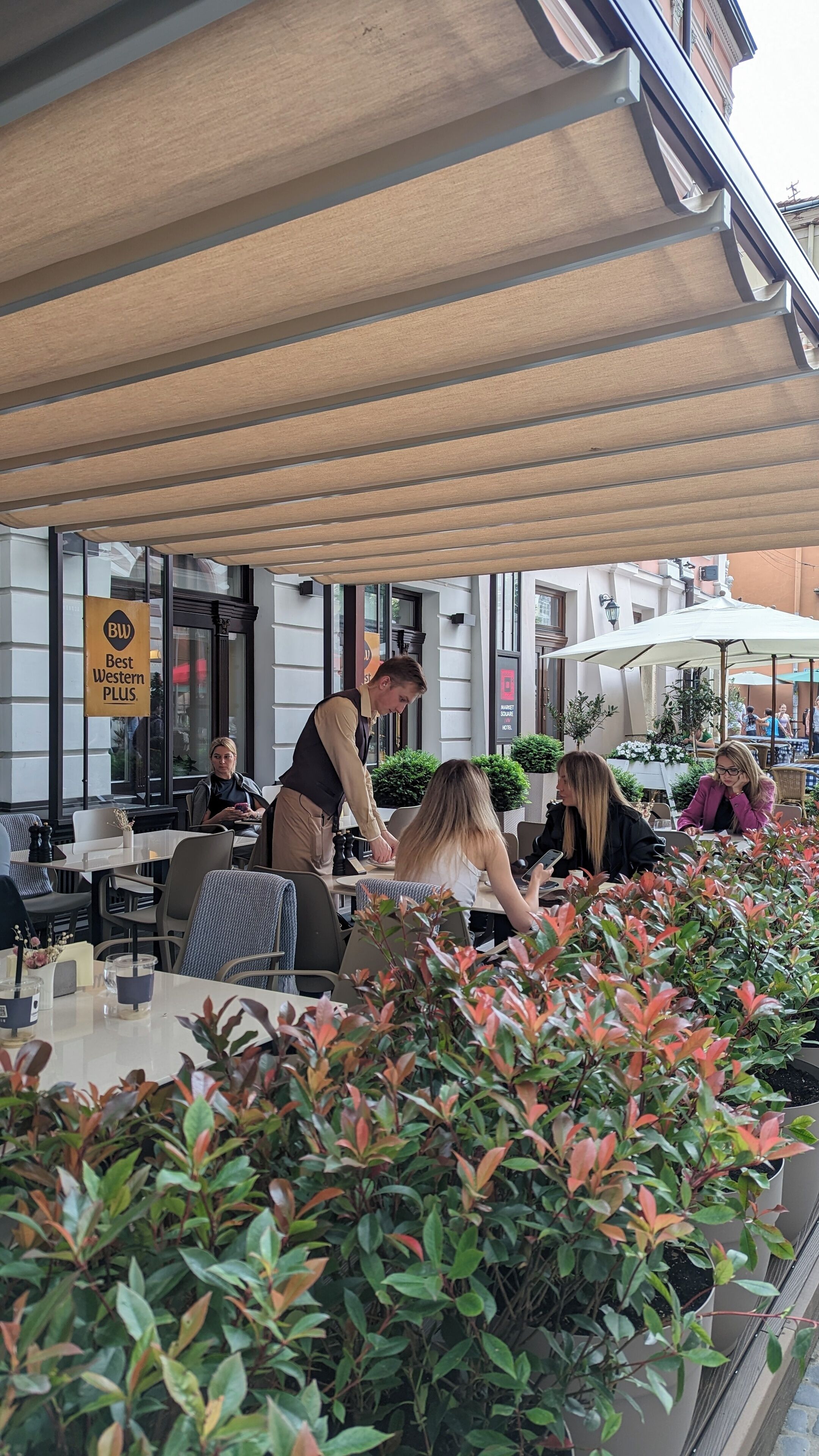 Un espace caf é en plein air avec des tables et des chaises, un serveur qui s’occupe d’un client, et une enseigne indiquant l’hôtel « Best Western PLUS ».